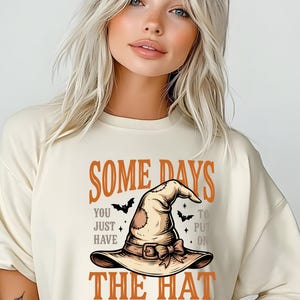 May include: A cream-coloured t-shirt featuring a graphic of a tan witch hat with a buckle and bow. The text reads: SOME DAYS YOU JUST HAVE TO PUT ON THE HAT AND REMIND THEM WHO THEY'RE DEALING WITH. Small black bats and stars surround the central hat illustration.