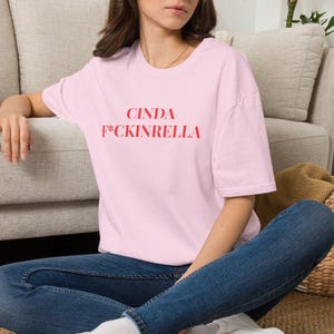 May include: A light pink short-sleeved t-shirt featuring the red text CINDA F*CKINRELLA printed on the chest. The garment has a crew neckline and a relaxed fit, paired with dark blue denim jeans.