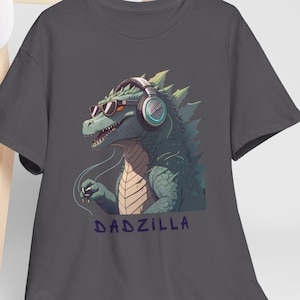 May include: Dark grey t-shirt with a green Godzilla-style creature wearing headphones and sunglasses. The creature has a detailed design with spikes and scales. The text "DADZILLA" is printed below the graphic in blue.