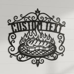 Puede incluir: Arte de pared de metal negro con un dise&ntilde;o de parrilla con llamas y un filete. Las palabras "CUSTOM TEXT" se muestran encima de la parrilla en una fuente decorativa. El arte est&aacute; montado en una pared blanca.
