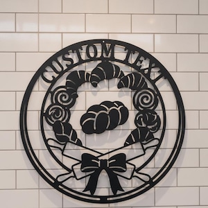 Könnte beinhalten: Ein kreisförmiges Wand-Schild aus schwarzem Metall mit dem Text CUSTOM TEXT am oberen Rand. Das Design zeigt einen zentralen Zopf, umgeben von verschiedenen Backwaren, zwei gekreuzten Nudelhölzern und einer dekorativen Schleife unten.