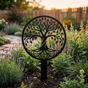 Op de afbeelding: Een zwarte metalen tuinpaal met een levensboomontwerp in een cirkelvormig frame. De boom heeft gedetailleerde bladeren en wortels. De paal staat in een tuin met diverse planten en bloemen, met een houten hek op de achtergrond.