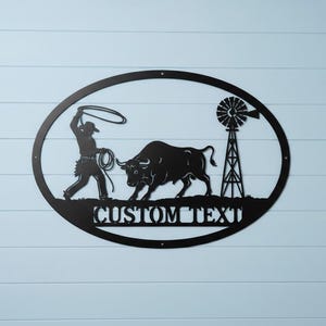 Op de afbeelding: Zwart metalen wandkunstwerk met een cowboy die een stier vangt met een lasso, met een windmolen op de achtergrond. Het ontwerp is geplaatst in een ovale lijst, met de tekst "CUSTOM TEXT" onderaan. Gemonteerd op een lichtblauwe, horizontaal beklede muur.