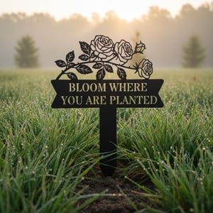 May include: Black metal garden stake with a floral design and the text "BLOOM WHERE YOU ARE PLANTED". The stake features a decorative rose branch and is placed in a field of green grass.