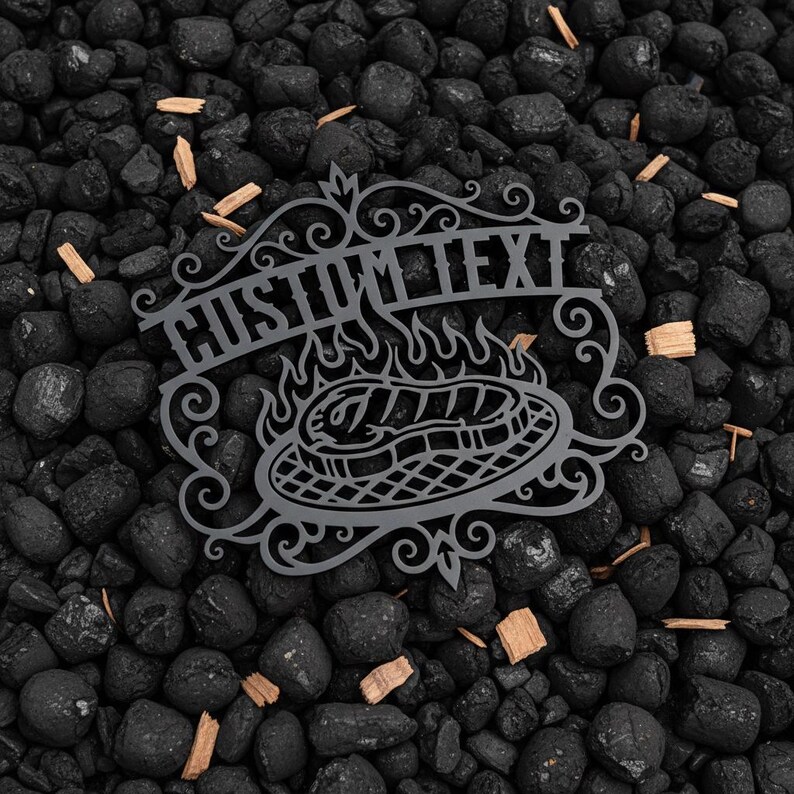 Puede incluir: Un letrero met&aacute;lico gris con la inscripci&oacute;n "CUSTOM TEXT" sobre una parrilla con llamas y un filete. El letrero est&aacute; rodeado de adornos decorativos. El letrero est&aacute; colocado sobre un lecho de rocas negras y virutas de madera.