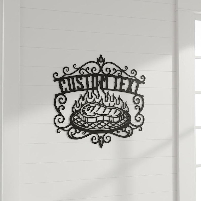 Puede incluir: Arte de pared de metal negro con un dise&ntilde;o de parrilla con llamas y un filete. Las palabras "CUSTOM TEXT" se muestran encima de la parrilla en una fuente decorativa. El arte est&aacute; montado en una pared blanca.