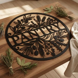 Op de afbeelding: Zwart metalen voorraadkastbord met een cirkelvormig ontwerp met het woord 'PANTRY' in grote letters, omgeven door olijftakken, tarwe en andere botanische elementen. Het bord staat op een houten oppervlak.