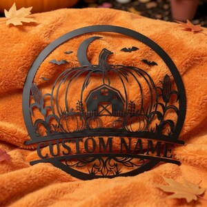 Pode incluir: Uma placa de parede circular em metal preto com um design recortado de uma abóbora grande, um celeiro, pés de milho e morcegos sob uma lua crescente. A seção inferior inclui uma barra horizontal com o texto CUSTOM NAME em fonte serifada.