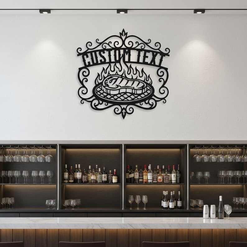 Puede incluir: Arte mural de metal negro con un dise&ntilde;o de parrilla con llamas y un filete. Las palabras "CUSTOM TEXT" se muestran encima de la parrilla. La obra de arte est&aacute; montada en una pared blanca sobre una barra.