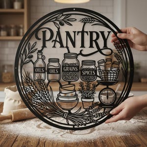 Könnte beinhalten: Schwarzes Metall-Vorratskammer-Schild mit dem Wort "Pantry" in eleganter Schrift. Das Schild zeigt ausgeschnittene Bilder von Gläsern mit der Aufschrift "Oils", "Grains" und "Spices" sowie einen Korb, eine Waage und Weizenähren. Das Schild wird von Händen gehalten.