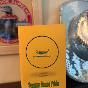 May include: A yellow booklet titled "Swamp Queer Chronicle" with a green alligator illustration inside a black circle. The text "First Edition March 2026" and "Swamp Queer Pride" are also visible. A framed image and a sequined hat are in the background.