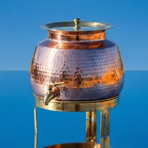 May include: A copper water dispenser with a hammered texture, brass accents, and a spigot. The dispenser sits on a brass stand, reflecting in the blue surface below. A cloud floats in the blue sky above.