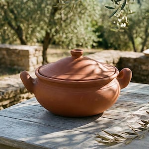 May include: A terracotta cooking pot with a lid and two handles sits on a weathered wooden table. The pot is a warm reddish-brown colour. The text "SLOW LIVING" is displayed in gold lettering at the bottom of the image. Olive branches are in the background.
