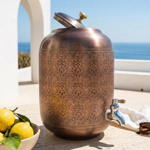May include: A copper-colored beverage dispenser with an ornate floral pattern. The lid is slightly ajar, revealing a gold-colored handle. A silver spigot is visible on the side. A bowl of lemons sits nearby. Sunglasses rest on the surface.