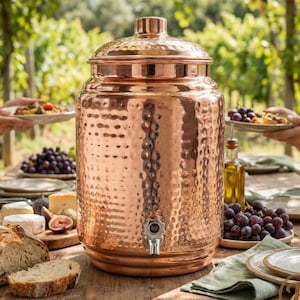 May include: A large, hammered copper drinks dispenser with a tap and lid. The dispenser is on a wooden table with food and drinks, including grapes, bread, and cheese. The background shows a vineyard.