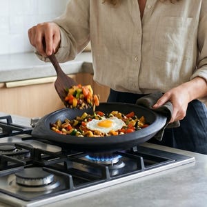 May include: A woman cooking in a modern kitchen, smiling while using a wooden spatula to serve a colorful vegetable and egg dish from a black skillet. The stove is on, with a blue flame visible. The kitchen has light wood cabinets and a large window.