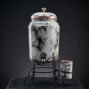 Puede incluir: Un dispensador de bebidas con diseño de mármol, borde cobrizo y tapa dorada. El dispensador está sobre un soporte de metal negro con una taza a juego. El texto "THE ART OF HYDRATION" está en la parte inferior.