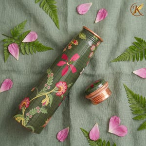 May include: A green water bottle with a floral design in pink, yellow, and white. The bottle has a copper-colored rim and a matching lid. Ferns and pink flower petals surround the bottle on a green cloth.