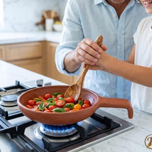 Op de afbeelding: Een terracotta pan met een handvat staat op een gasfornuis. De pan bevat rode en gele kerstomaatjes en groene basilicumblaadjes. Een houten lepel roert de inhoud. Een persoon en een kind glimlachen tijdens het koken.