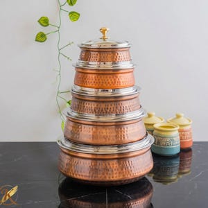 Puede incluir: Una pila de ollas de cocina de color cobre con bordes y tapas plateadas. Las ollas tienen una textura martillada y están dispuestas de mayor a menor. Pequeños tarros de cerámica en amarillo, azul y naranja están en el fondo.