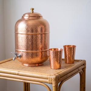 Puede incluir: Un dispensador de agua cilíndrico grande de cobre martillado con tapa a juego y un grifo de metal plateado, acompañado de dos vasos de cobre martillado a juego. El conjunto presenta un acabado metálico pulido y reflectante.