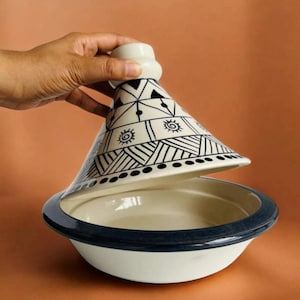 May include: A ceramic tagine with a conical lid featuring black geometric patterns, including triangles, sunbursts, and lines on a white background. The base is a wide, shallow bowl with a dark blue rim, designed for cooking and serving food.