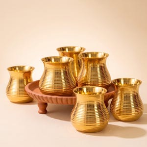 May include: A set of six gold-colored metal cups with a horizontal ribbed texture and a flared rim, arranged on a round, terracotta-colored pedestal tray with a scalloped edge. The cups have a rounded body shape and a polished, reflective finish.
