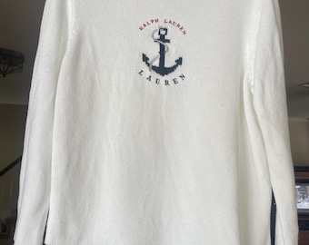 May include: White knit sweater with a navy blue anchor design and the words "RALPH LAUREN" embroidered above and below. The bottom hem features three navy blue stripes. The sweater is hanging on a black hanger.