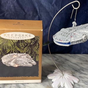 Puede incluir: Adorno Hallmark Keepsake del Halcón Milenario de Star Wars. La nave espacial plateada tiene detalles azules y rojos y cuelga de un soporte plateado con una base blanca. El adorno está junto a su caja original.