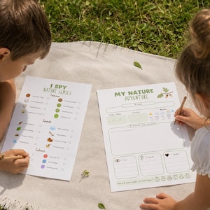 May include: Two children are seated on a blanket, each with a nature-themed activity sheet. One sheet is titled "I Spy Nature Senses" and the other is "My Nature Adventure." The children are using pencils to fill out the sheets.