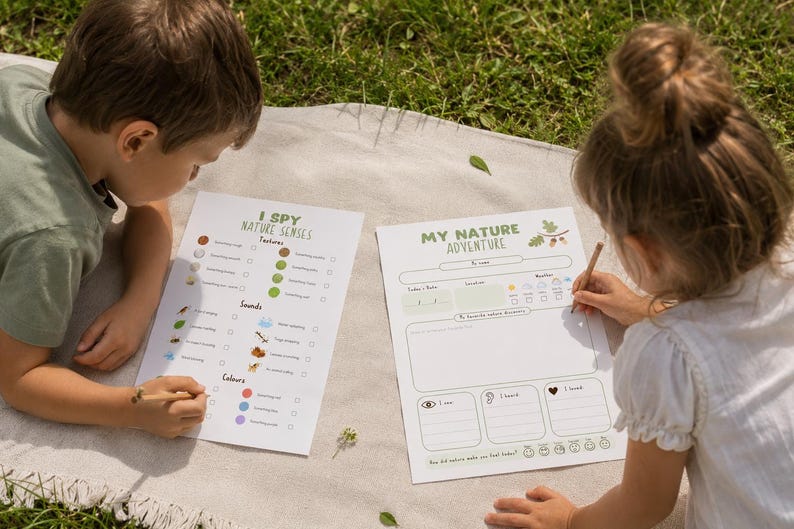 May include: Two children are seated on a blanket, each with a nature-themed activity sheet. One sheet is titled "I Spy Nature Senses" and the other is "My Nature Adventure." The children are using pencils to fill out the sheets.