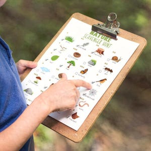 May include: A clipboard with a "Nature Scavenger Hunt" checklist. The checklist includes illustrations of natural items like leaves, insects, and a bird. The clipboard has a metal clip and is held by a person wearing a blue shirt.