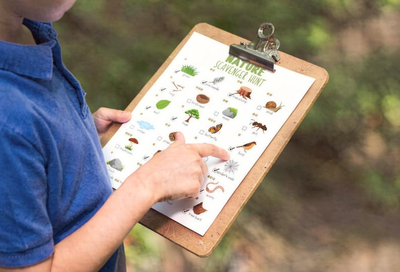 May include: A clipboard with a "Nature Scavenger Hunt" checklist. The checklist includes illustrations of natural items like leaves, insects, and a bird. The clipboard has a metal clip and is held by a person wearing a blue shirt.
