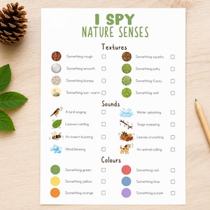 May include: A printed "I Spy Nature Senses" activity sheet with illustrations and check boxes. The sheet includes categories for textures, sounds, and colors. A pinecone, leaves, and a pencil are on the wooden surface.
