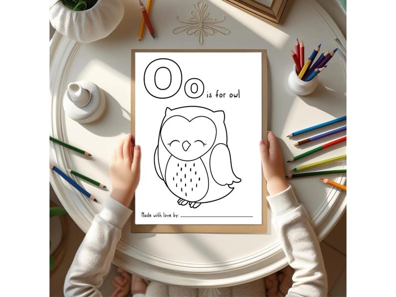May include: A coloring page with the letter "O" and the phrase "is for owl" above a cartoon owl illustration. The page is held by hands, surrounded by colored pencils and art supplies.