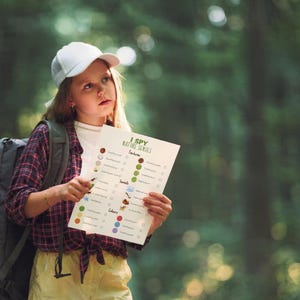 May include: A young person wearing a white cap, backpack, and plaid shirt holds an "I Spy Nature Series" activity sheet in a forest setting. The sheet features colorful illustrations and check boxes for nature-themed items.