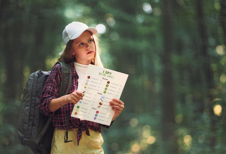 May include: A young person wearing a white cap, backpack, and plaid shirt holds an "I Spy Nature Series" activity sheet in a forest setting. The sheet features colorful illustrations and check boxes for nature-themed items.