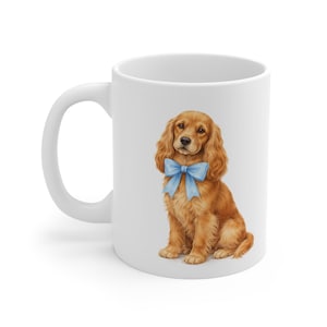 Könnte beinhalten: Weiße Keramiktasse mit einer detaillierten Illustration eines goldenen Cocker Spaniel-Welpen mit einer hellblauen Schleife. Der Hund sitzt mit flauschigem Fell. Die Tasse hat einen gebogenen Griff.