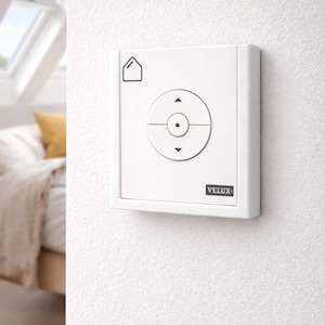 Peut inclure: Un panneau de commande mural blanc avec un cadran de commande circulaire et des flèches directionnelles. Le panneau comporte une icône de maison et la marque "VELUX" est visible. Le panneau est monté sur un mur blanc texturé.