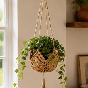 Pode incluir: Um suporte de plantas suspenso em macramê com uma planta verde em vaso. O suporte é feito de corda trançada de cor bege, com uma borla decorativa na parte inferior. A planta tem folhas verdes que caem pelas laterais.