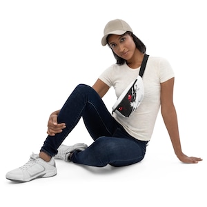 May include: White fanny pack with a black and red graphic design. The bag has a black adjustable strap and a zipper closure. The fanny pack is worn across the body. The person in the image is wearing a white t-shirt, blue jeans, and a beige baseball cap.