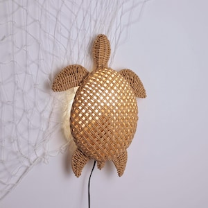 Puede incluir: Una lámpara de pared con forma de tortuga de ratán tejido. La lámpara tiene una superficie texturizada y se ilumina desde el interior, emitiendo un brillo cálido. La lámpara está fijada a una pared blanca con un cable de alimentación negro.