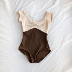 May include: A short-sleeved leotard featuring a dark brown lower section and a textured, cream-colored upper bodice with a V-neckline and a gathered center detail. The garment has a clean, fitted silhouette with a classic leg cut.