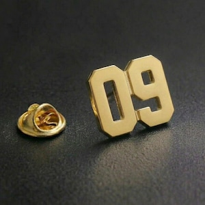 May include: A gold-coloured pin displaying the number '09' in a bold, blocky font. The pin has a classic design with a pin back for secure attachment. The pin is likely made of metal and has a shiny finish.