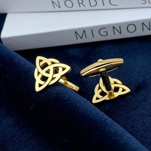 Könnte beinhalten: Ein Paar Manschettenknöpfe aus goldfarbenem Metall mit einem durchbrochenen Triquetra-Knotenmuster. Ein Manschettenknopf ist so positioniert, dass die Vorderseite mit dem verschlungenen Muster sichtbar ist, während der andere das Profil zeigt.