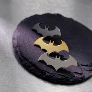 May include: Three metal bat symbols in black, gold, and silver are arranged on a dark, textured surface. The bat symbols are stylized with pointed wings and a central notch, showcasing a superhero theme.