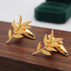 May include: A pair of gold-toned metal cufflinks featuring a polished finish. Each cufflink is shaped like a delicate botanical sprig with multiple pointed leaves extending from a central stem, attached to a smooth, rounded bullet-back closure.