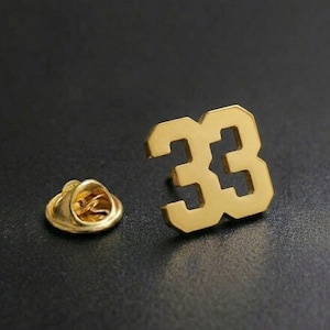 May include: A gold-colored lapel pin in the shape of the number 33. The pin has a classic design with a gold clutch back. The pin is shown on a dark background.