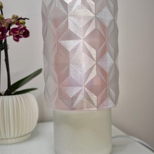 Può includere: Una lampada da tavolo geometrica bianca con base strutturata e paralume sfaccettato e iridescente. Il paralume ha un motivo a diamante. Un'orchidea in vaso è sullo sfondo.