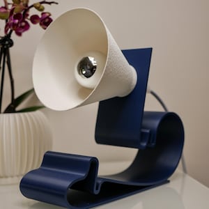 Può includere: Una moderna lampada da scrivania con un paralume bianco strutturato e una lampadina riflettente. La base è blu navy con un design curvo a forma di onda. La lampada è su una superficie bianca.